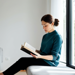 A professional adult with a serene and focused expression, sitting comfortably in a sunlit, minimalist living space. They are fully clothed in modest, appropriate attire and are deeply engrossed in reading a physical book, with no digital devices visible. The background is clean and uncluttered, featuring soft, natural light filtering through a large window, conveying an atmosphere of tranquility and mindful engagement. safe for work, appropriate content, fully clothed, professional, family-friendly, perfect anatomy, correct proportions, natural pose, well-formed hands, proper finger count, natural body proportions, professional photography, high quality.